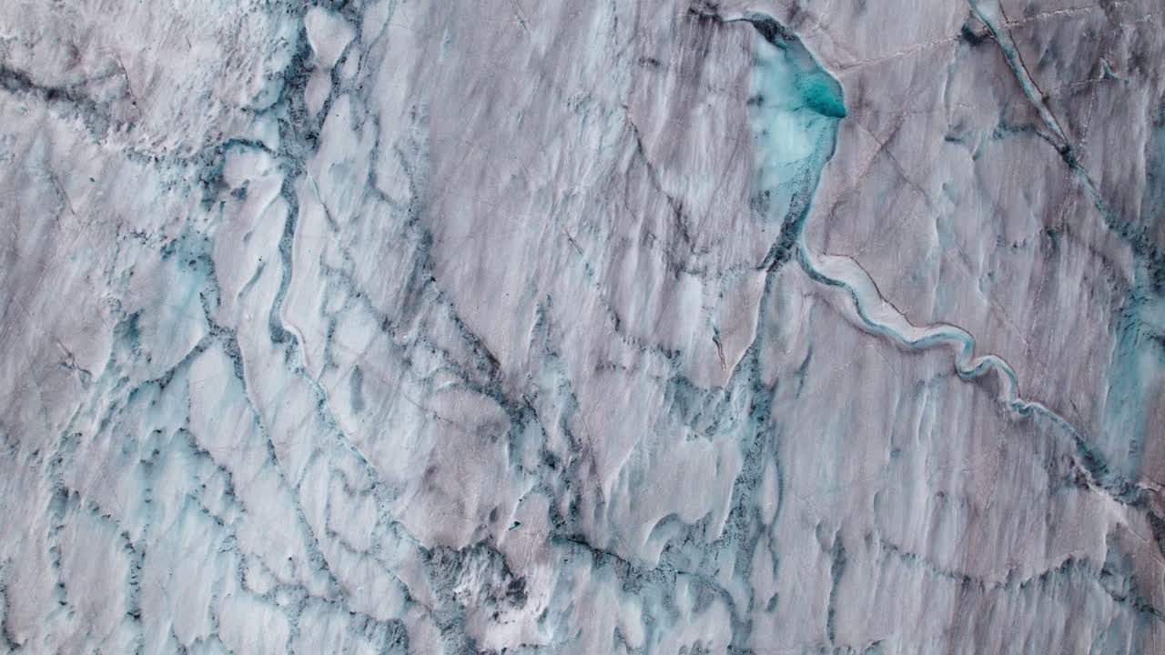 Vertikale 4K-Drohnenansicht der Oberfläche des Aletschgletschers, die Gletscherspaltennetze, Schmelzkanäle und gebrochene Eismuster zeigt, die durch Spannung, Fließdynamik und saisonales Schmelzen in den Schweizer Alpen entstanden sind