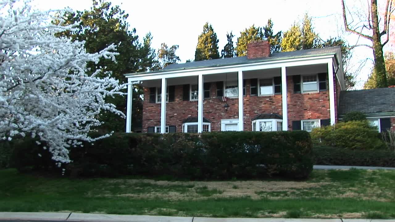 카메라는 앞마당 가장자리에 서 있는 꽃나무에서 하얀 기둥이 있는 붉은 벽돌집으로 확대됩니다.