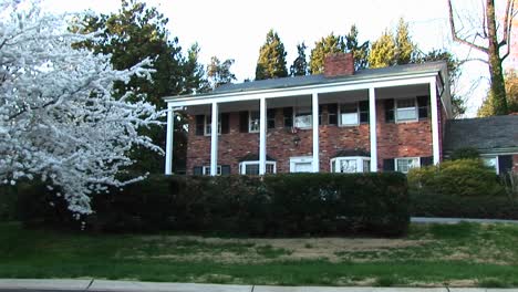 the camera zooms in from a flowering tree stands at edge of the front yard to the redbrick house with white pillars