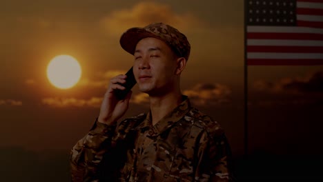 close up of side view asian man soldier talking on smartphone and looking around while standing with flag of the united states, sunset time