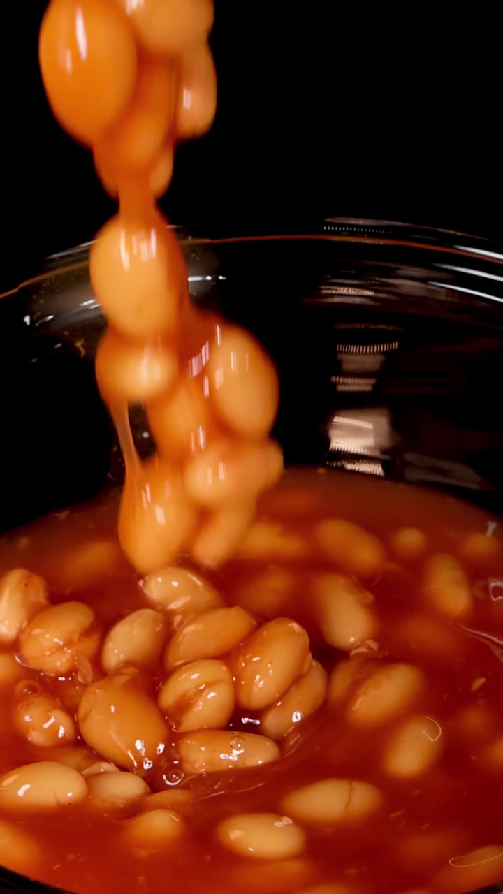 White beans in tomato sauce fall into a clear glass bowl against a black background, captured in slow motion with dramatic studio lighting and close-up framing