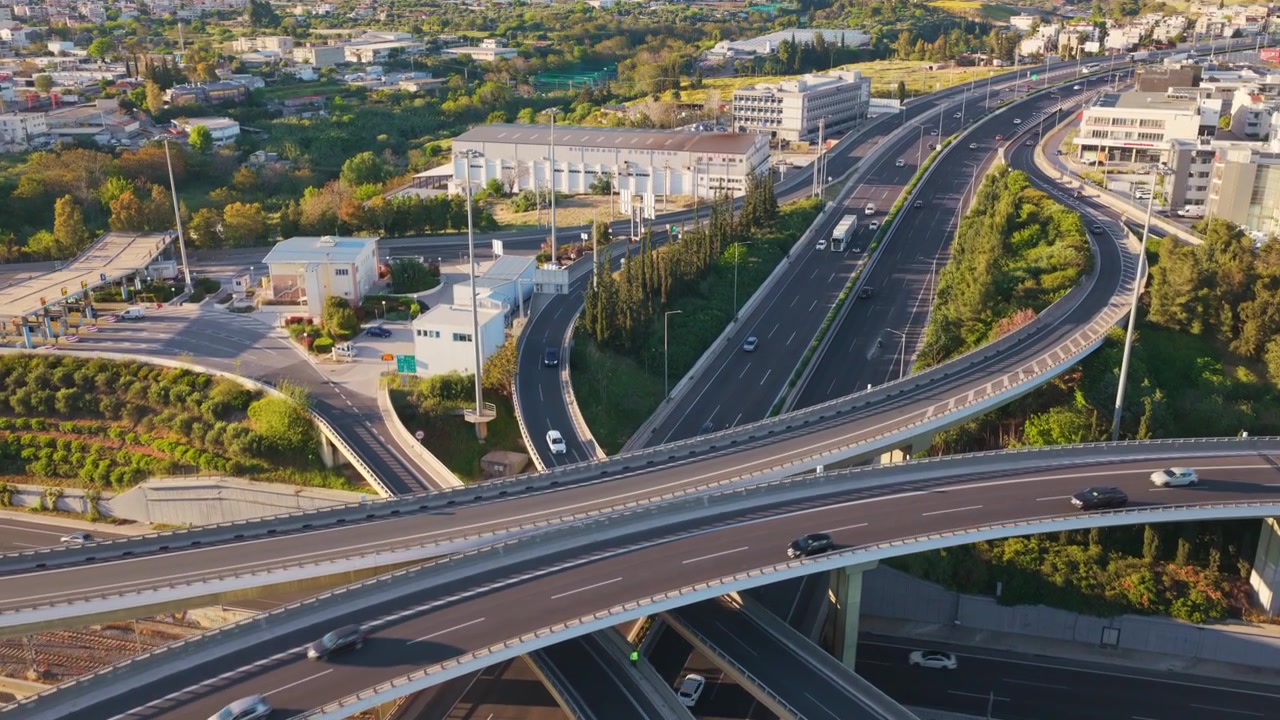 Ilmakuvaa monikerroksisesta moottoritieliittymästä, jossa autot ja kuorma-autot ajavat kaarevia ylikulkusiltoja ja ramppeja pitkin, kaupunki-infrastruktuurin ja vihreiden puiden ympäröimänä kauniin auringonlaskun aikaan.