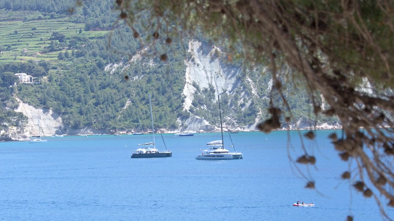 Inquadratura ampia e statica di yacht bianchi ormeggiati in acque calme e turchesi vicino a scogliere calcaree costiere.
