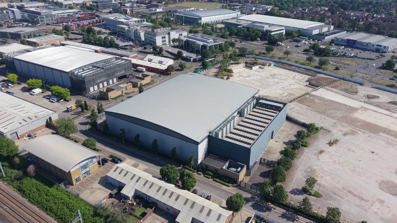 Drone view over a large data center building and surrounding industrial complex. Many warehouses, parked cars, and active railway tracks are visible from above during a clear daytime.