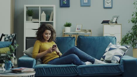 Attractive-Young-Curly-Lady-Sitting-On-The-Sofa-And-Resting-While-Tapping,-Scrolling-And-Texting-On-The-Smartphone-At-Home