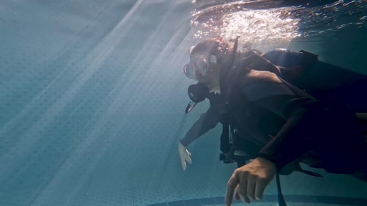 Side underwater shot of a woman in scuba gear in a pool, she turns to the camera and waves in the sun's glare, then continues swimming