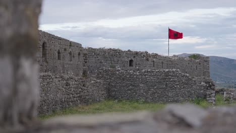 gray medieval stone walls with arched windows and a red albanian flag waving above rozafa castle, albania