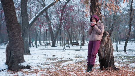 dama con traje de invierno descansando contra un árbol comiendo algo en un parque al aire libre durante el invierno, suelo nevado, fondo del parque, disfrutando de la comida, actividad casual de invierno