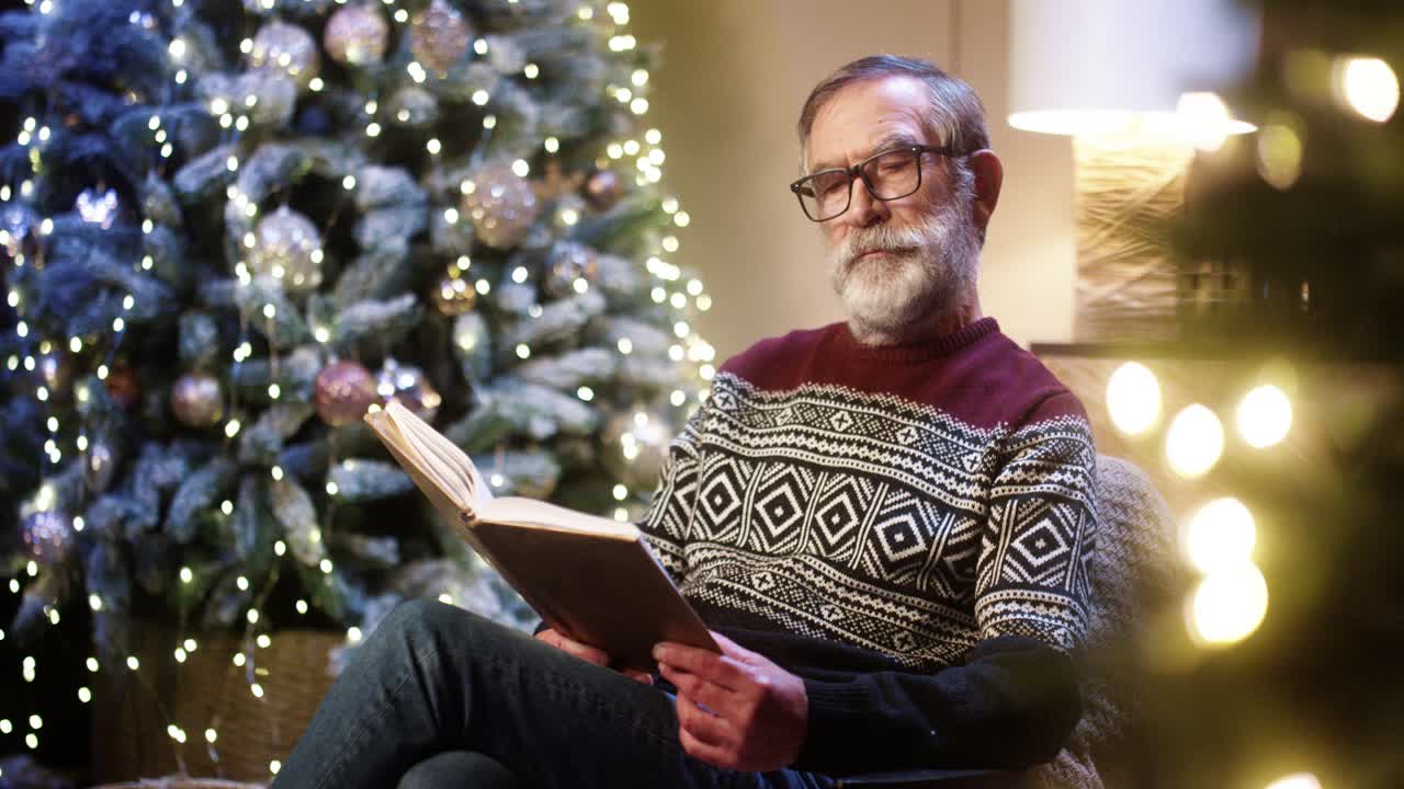 retrato de un viejo y agradable abuelo jubilado con gafas y un suéter de navidad leyendo un libro mientras se sienta en una habitación decorada cerca de un árbol de navidad resplandeciente