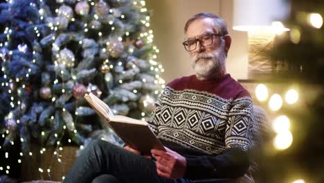 portrait of old nice male pensioner grandpa in glasses and christmas sweater reading book while sitting in decorated room near glowing christmas tree