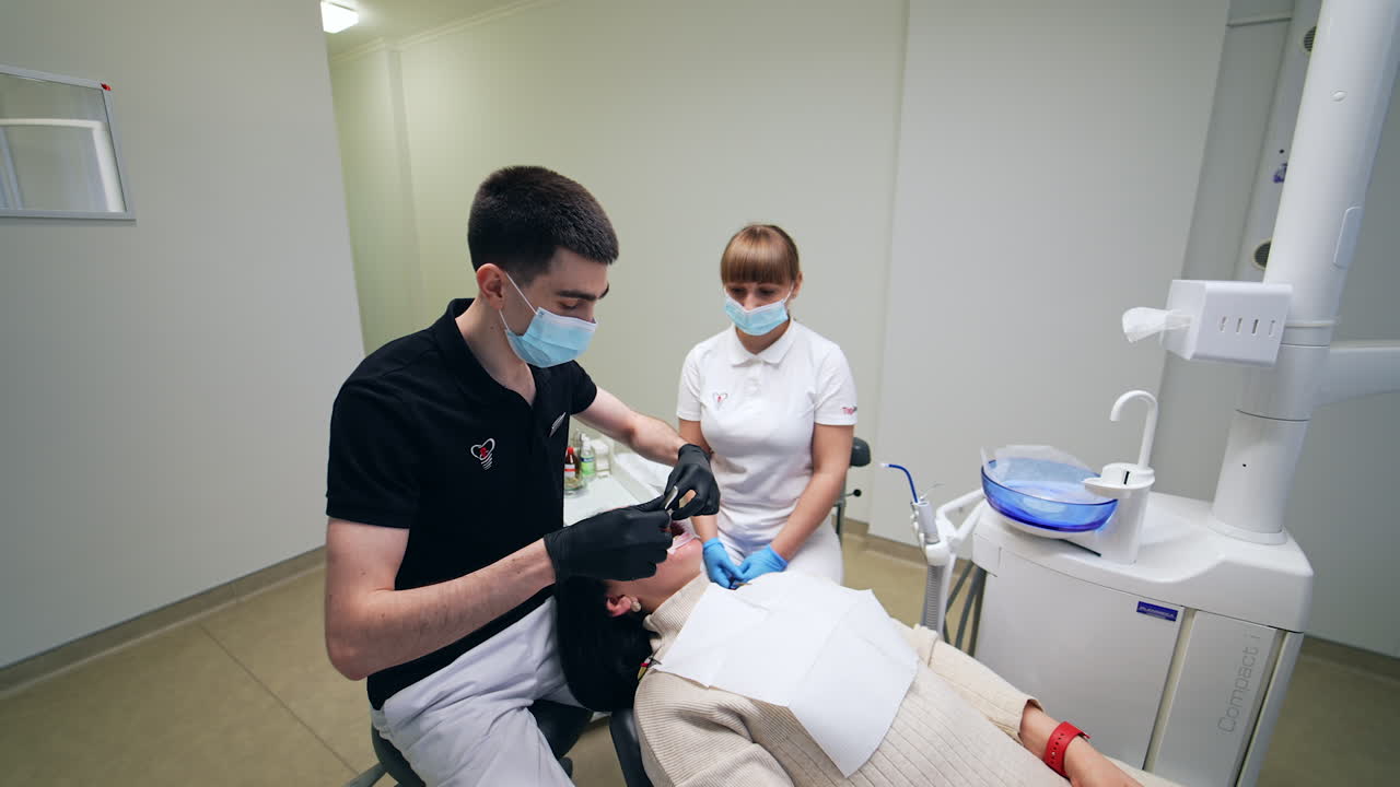 Dentist in black t-shirt working in the light spacious cabinet. Doctor uses metal tool in his work. Female nurse sitting at backdrop.