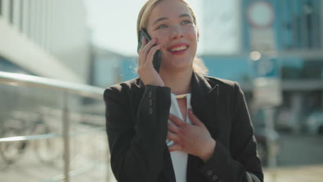 Cheerful lecturer in professional outfit walking confidently while talking on phone with friend, smiling with joy as person approaches from behind in urban setting with glass building background