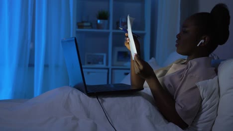 Afrikanische-Frau-Mit-Laptop,-Die-Nachts-Im-Bett-Arbeitet.Technologie,-Remote-Job-Und-People-Konzept,-Glücklich-Lächelnde-Junge-Afroamerikanische-Frau-Mit-Kabellosen-Kopfhörern,-Laptop-Computer-Und-Papieren,-Die-Nachts-Zu-Hause-Im-Bett-Arbeitet