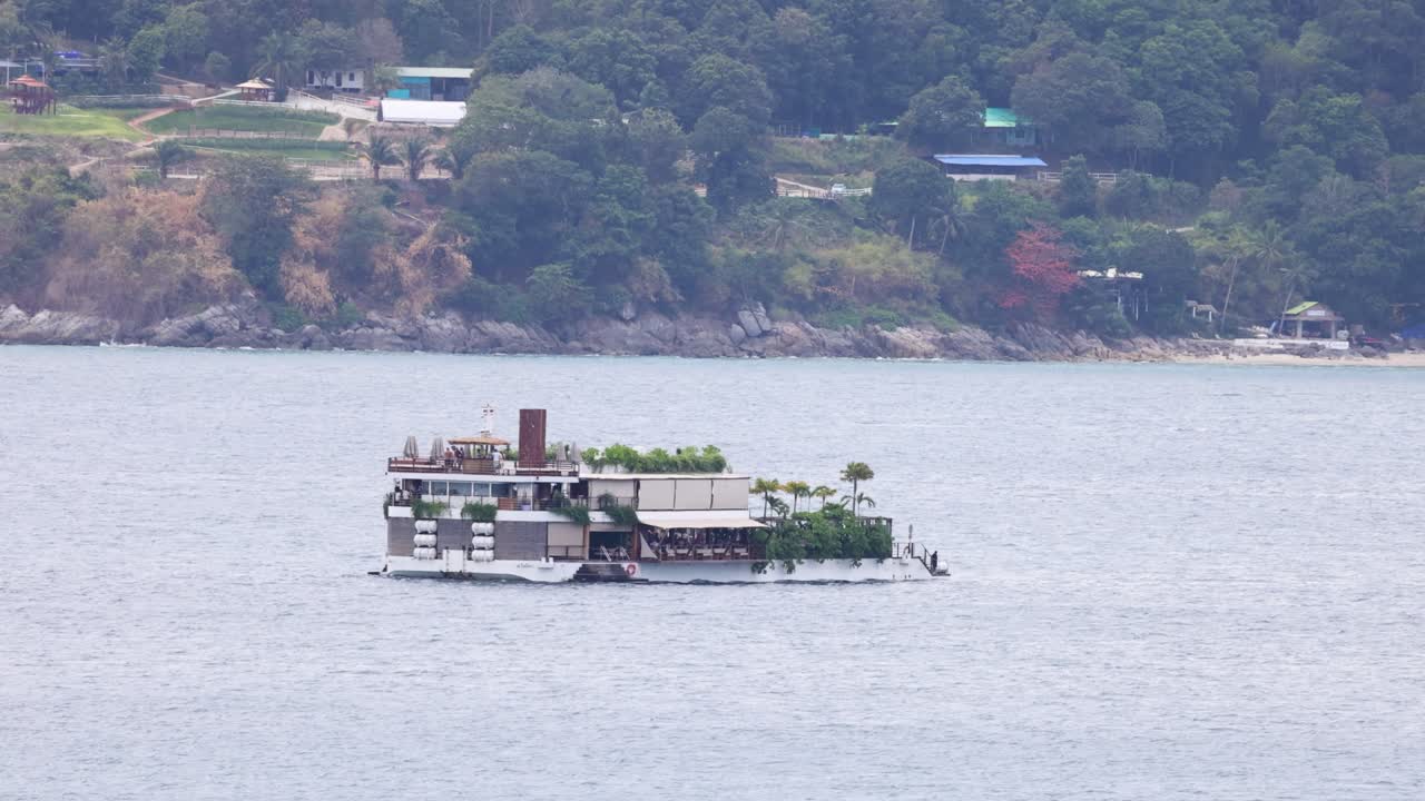 푸켓의 평온한 물을 가로질러 부드럽게 떠다니는 레스토랑, 울창한 초록색과 먼 언덕으로 둘러싸여
