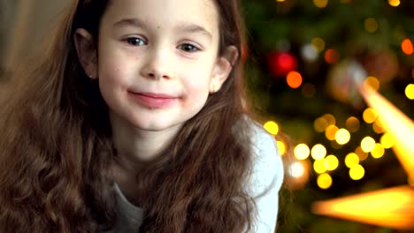 portrait of happy little girl sitting near christmas tree on new year's eve in decorated room. new year and holidays with kids concept. enjoying festive holiday celebration.