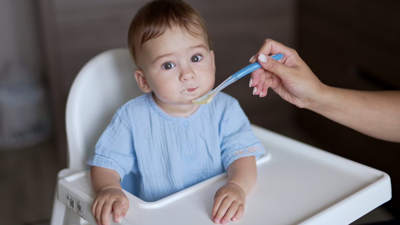 Sweet kid in blue shirt sits at feeding table. Mother giving a full spoon to her child. Close up. Blurred backdrop.