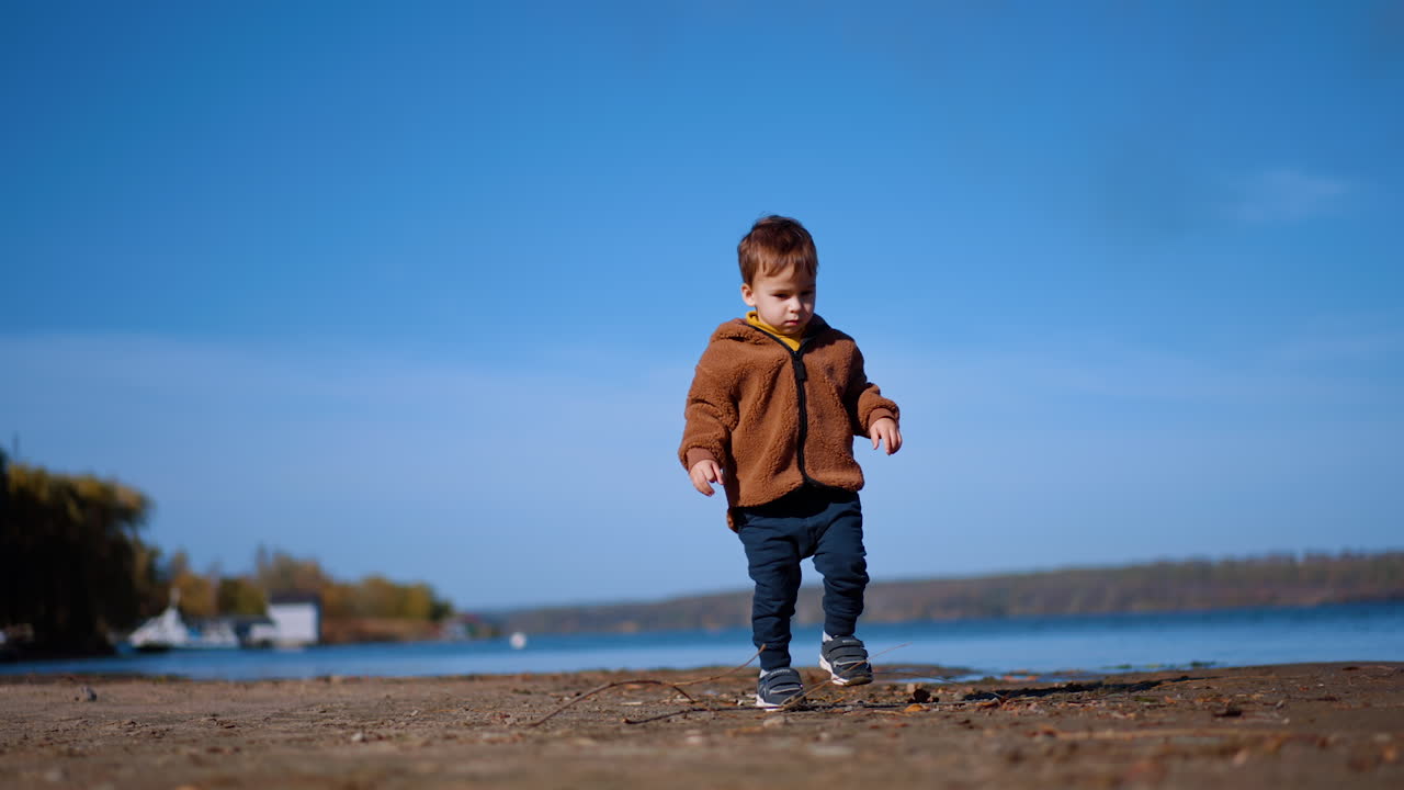 Anak lucu berjalan di tepi air mencari batu. Bayi laki-laki menyentuh ranting lalu melanjutkan perjalanan. Pandangan sudut rendah.