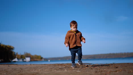 cute kid walks by the waterfront looking for the stones. baby boy touches a twig and then continues to go. low angle view.