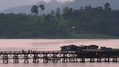 Mon-Bridge-and-a-longboat-followed-speeding-behind-the-structure-while-people-are-walking-on-the-bridge,-silhouetting-as-it-was-getting-dark,-in-slow-motion