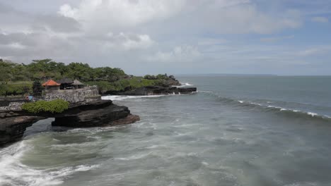 Aerial-skims-shorebreak-waves-toward-cliffs-of-Tanah-Lot-temple,-Bali