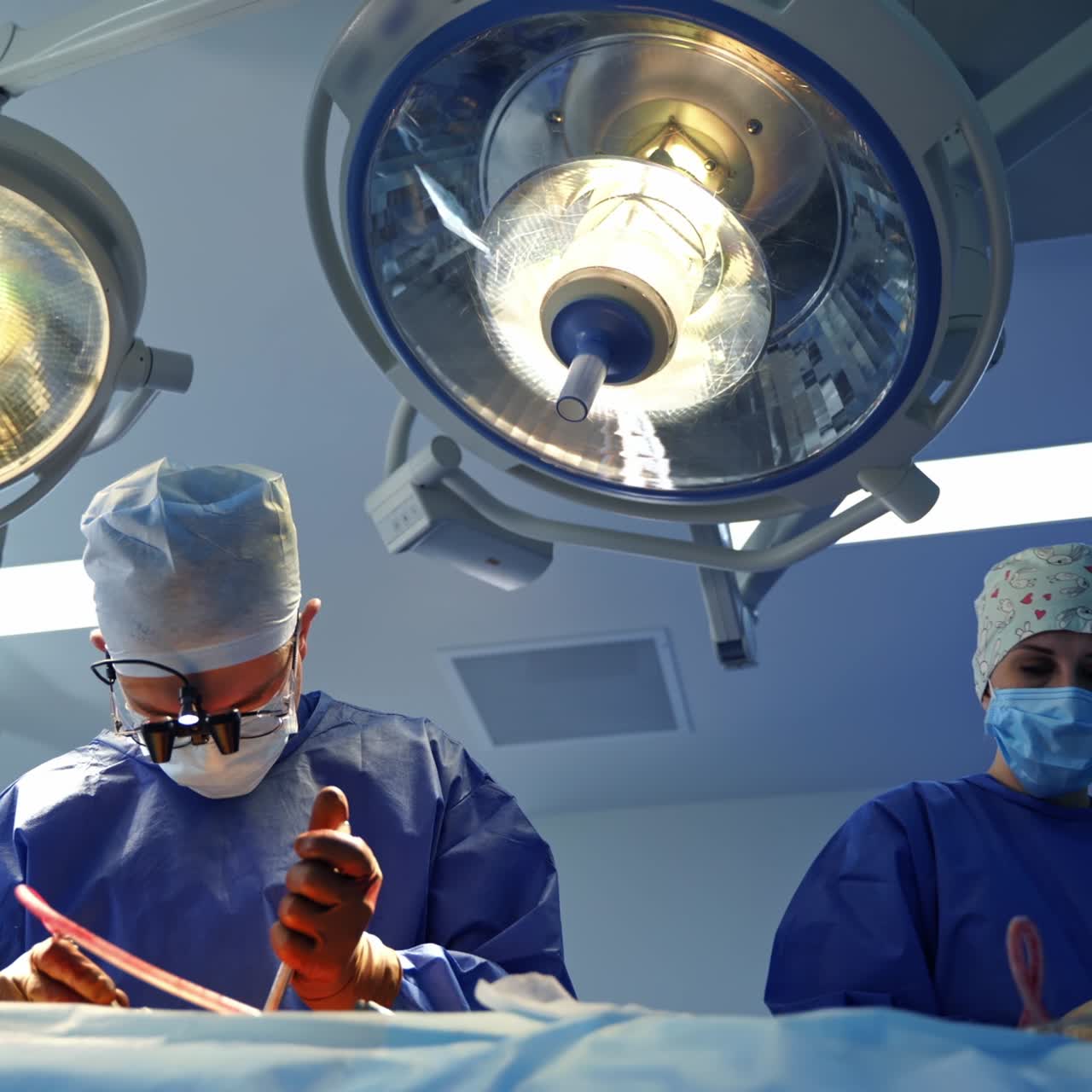 Doctor and nurse in masks operate patient. Medical workers perform a surgery under the bright medical lamps in the operating room. View from below