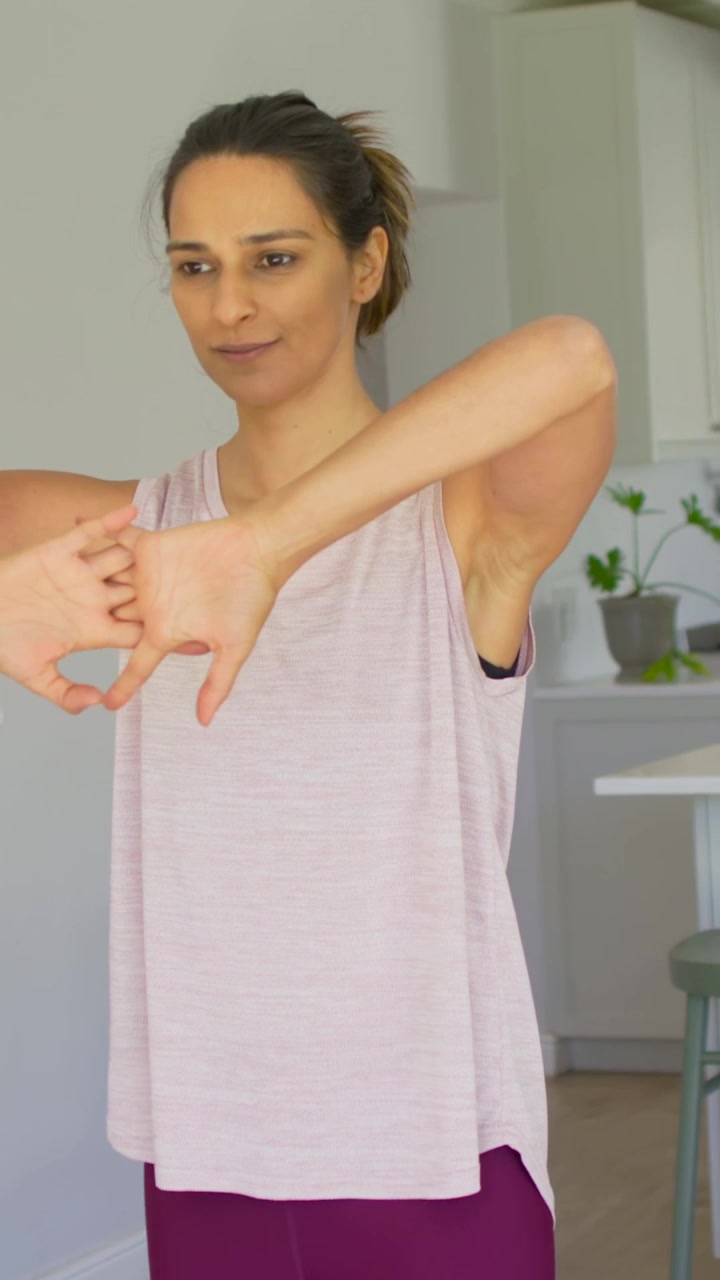 Video vertical: Mujer comenzando calentamiento de brazos en la cocina, con camiseta sin mangas rosa y leggings morados, junto a la encimera.