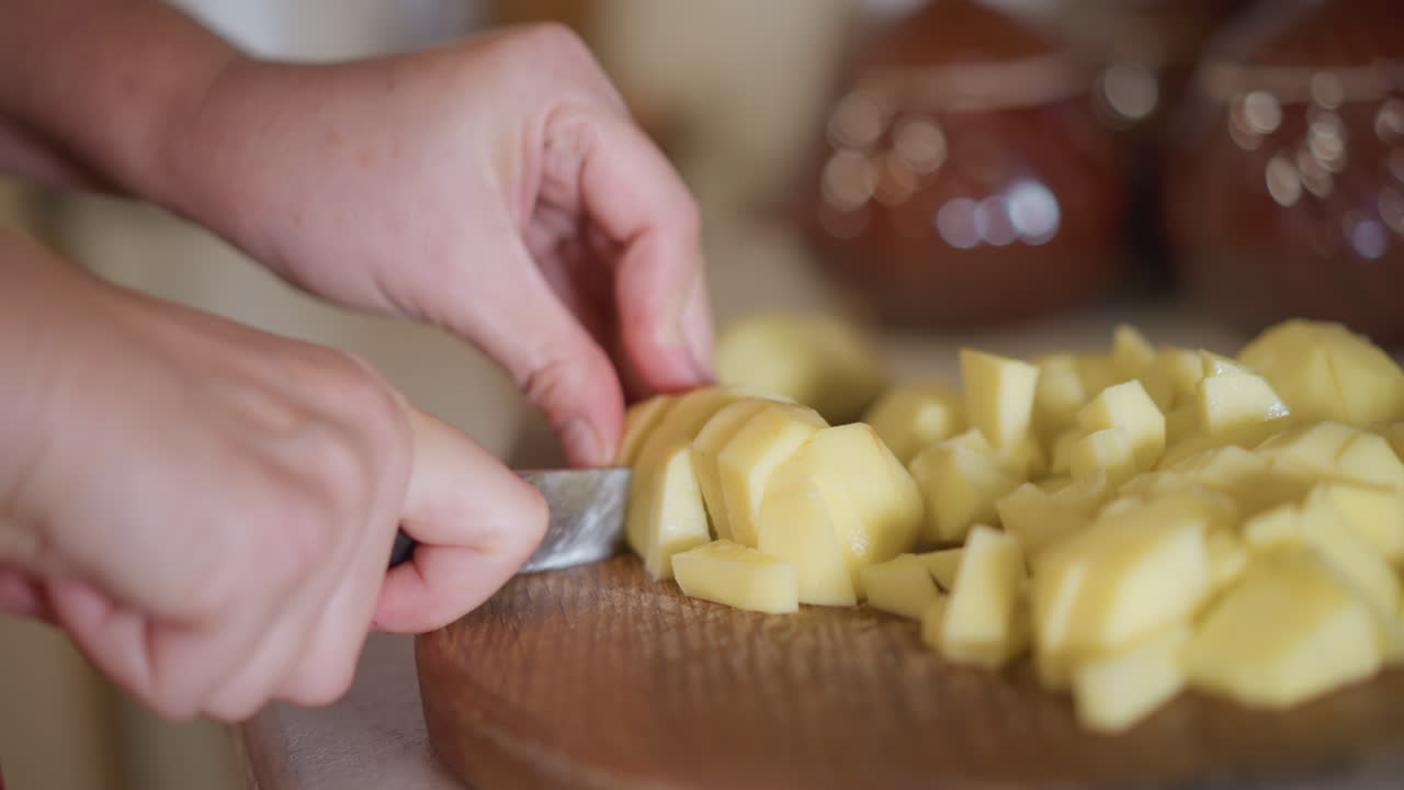 Chef casero picando patatas cerca de jarras de cerámica. Escena de cocina rústica con una persona cortando patatas rápidamente. Escena de cocina casera con una persona preparando patatas en una tabla de madera redonda.