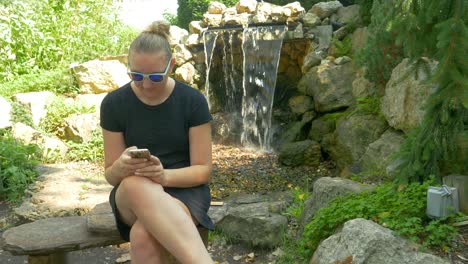 Frau-Lacht,-Lächelt-Und-Schüttelt-Den-Kopf,-Während-Sie-Neben-Einem-Kleinen-Wasserfall-Im-Park-Auf-Ihr-Smartphone-Schaut