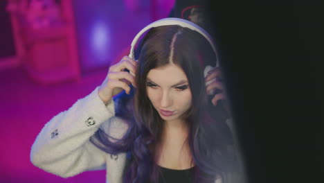 aerial view of young woman adjusting white headphones and resting hand on desk under pink purple ambient lighting at gaming live stream setup featuring dual monitors rgb keyboard glow