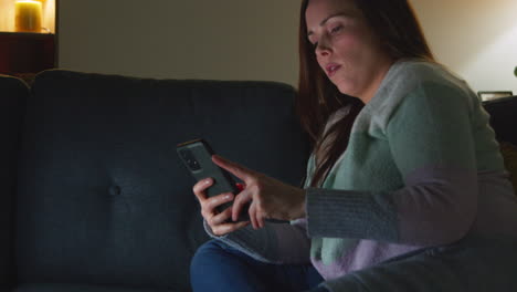 woman sitting on sofa at home at night streaming or watching movie or show on laptop and scrolling internet on phone 9