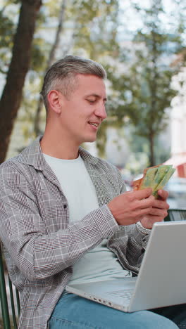 happy rich man using laptop winner victory holding cash money in euro banknotes sitting on chair