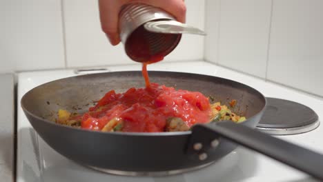 man pouring crushed tomatoes on fried vegetables in a frying pan