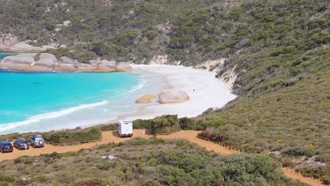 Drone-flying-over-carpark-at-Little-Beach-in-Albany,-Western-Australia-following-couple-walking-along-the-sand