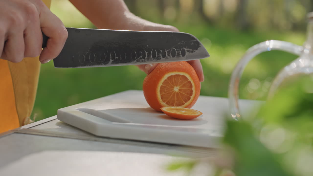Tavolo da campeggio soleggiato dove si affetta un'arancia fresca. Mani reggono un coltello su un tagliere bianco, la luce del sole mattutina fa brillare il succo sulla buccia. Un campeggiatore informale prepara uno spuntino salutare per la colazione in un allestimento all'aperto.