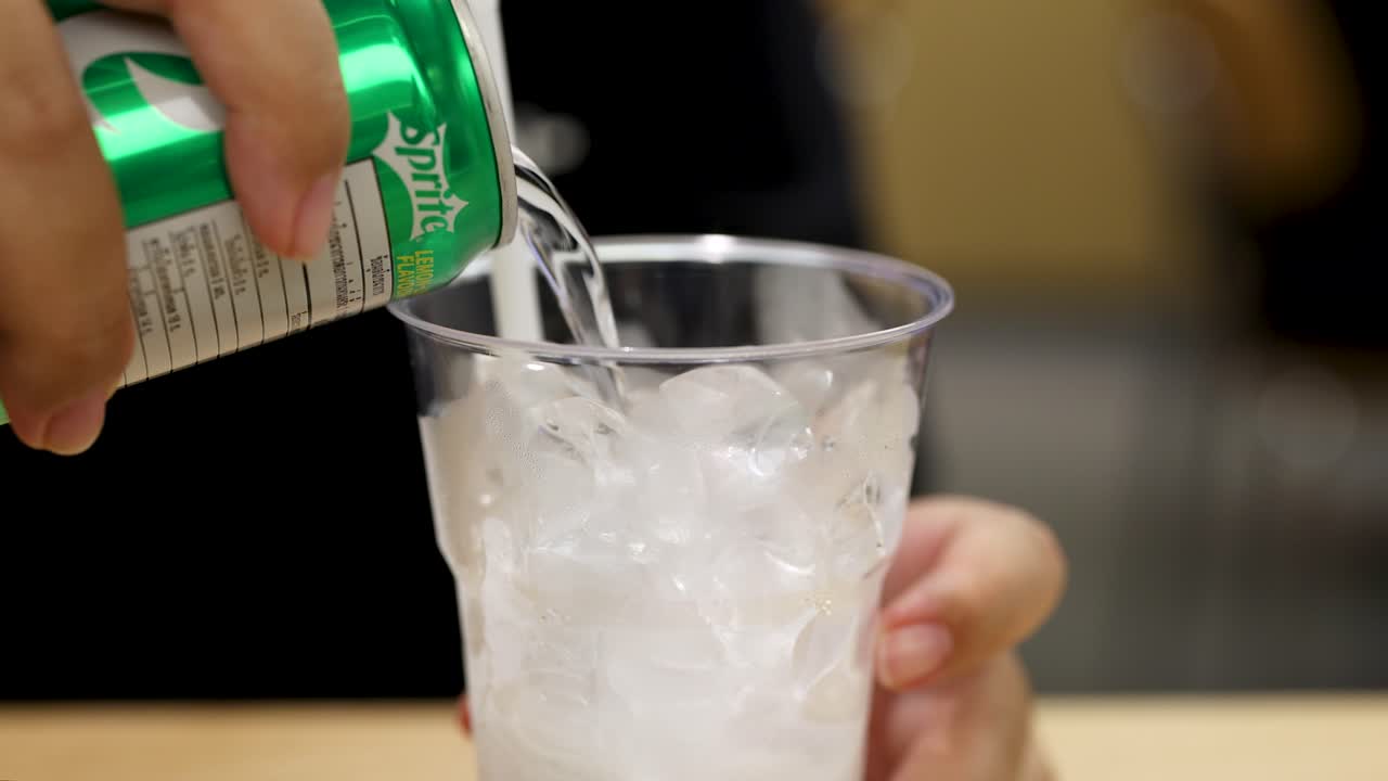 Hand pours carbonated soft drink into clear cup with ice, close-up, indoor, soft lighting