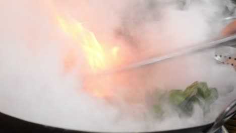 chef is stirring green vegetables in wok with flame and steam rising inside the kitchen