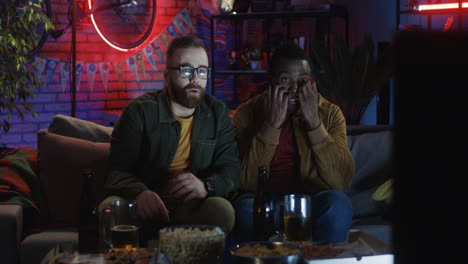 portrait shot of the two young men, caucasian and african american, watching horror movie on the sofa at home at night and being very scary.