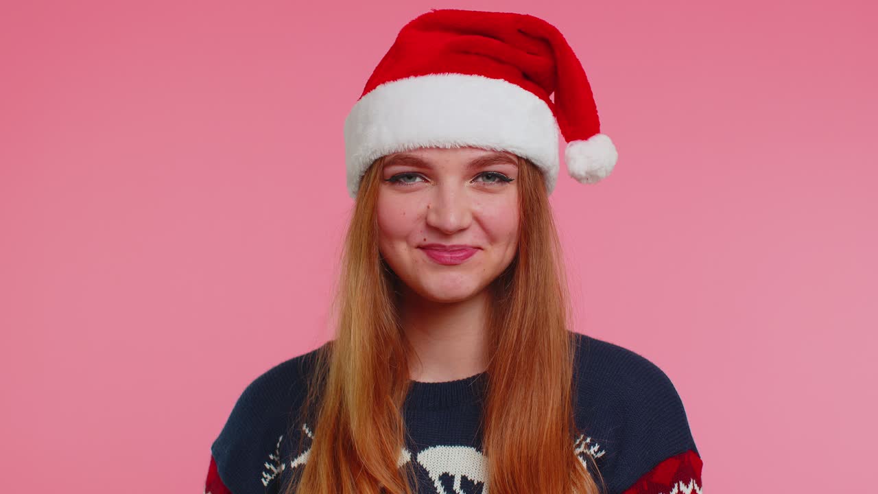 크리스마스 산타 스웨터에서 즐겁게 웃는 소녀는 카메라를 향해 금빛 반이는 콘페티를 불어넣습니다.