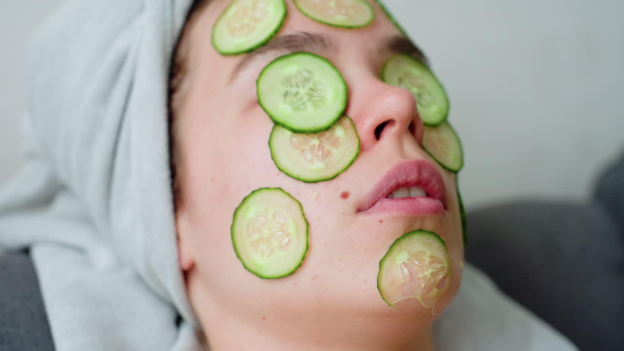 Close-up view of woman's face with cucumber slices placed on skin, relaxing facial treatment, hair wrapped in towel, soothing spa beauty routine, skin care, and self-care concept