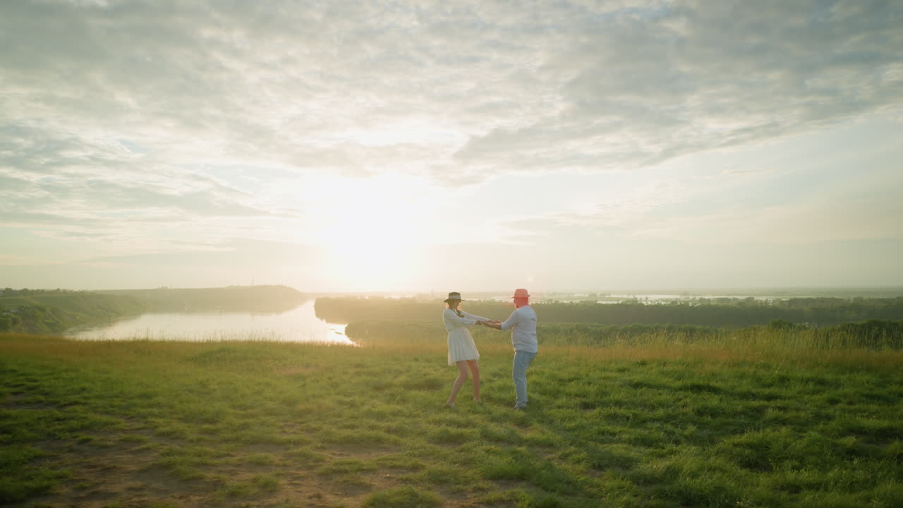 한 사랑하는 부부는 평온한 호수 의 잔디 언덕에서 해가 지는 동안 기게 회전합니다. 남편은  셔츠와 모자를 입고 색 드레스를 입은 아내를 포옹하여 순수한 로맨스의 순간을 포착합니다.