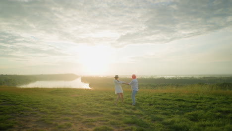 a loving couple spins joyfully on a grassy hill at sunset, beside a calm lake. the husband, in a white shirt and hat, embraces his wife in a flowing white dress, capturing a moment of pure romance