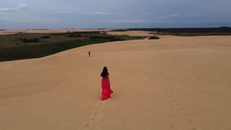 órbitas-Aéreas-Latina-Con-Vestido-Rojo-Paseando-Con-Perro-En-Una-Duna-De-Arena-De-Bolivia