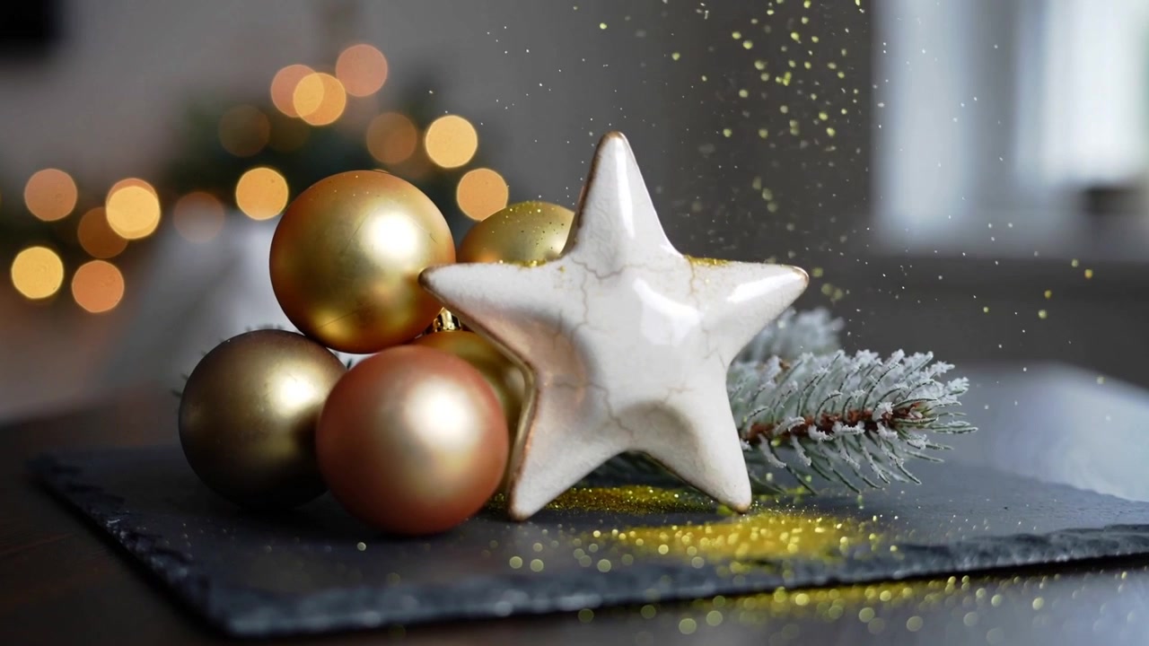 Stella di ceramica e palline decorative che ricevono glitter dorati che cadono su ardesia scura, con effetto bokeh.