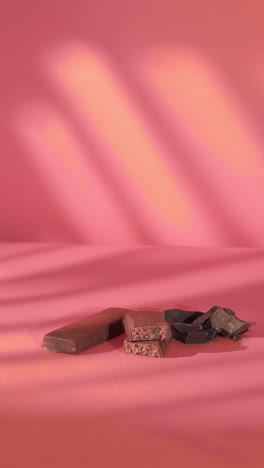 A hand reaches for a strawberry-flavored protein bar with a visible pink interior, stacked alongside chocolate bars and broken dark chocolate pieces on a soft pink background, diffused shadow lighting