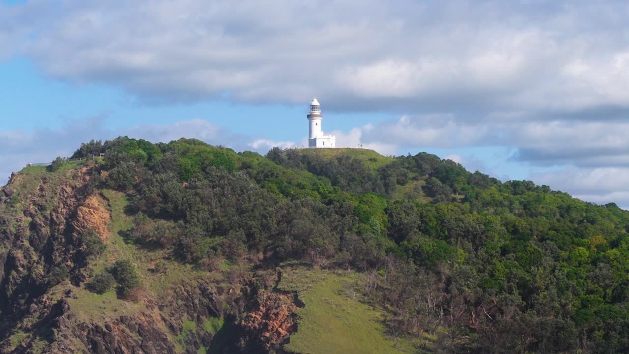 A lighthouse stands atop a lush, green hill under a partly cloudy sky in Byron Bay, Australia