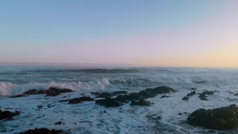 low flying drone shot flying over open ocean at sunset as waves rolls in and crashes on rocks with bamboo floating around