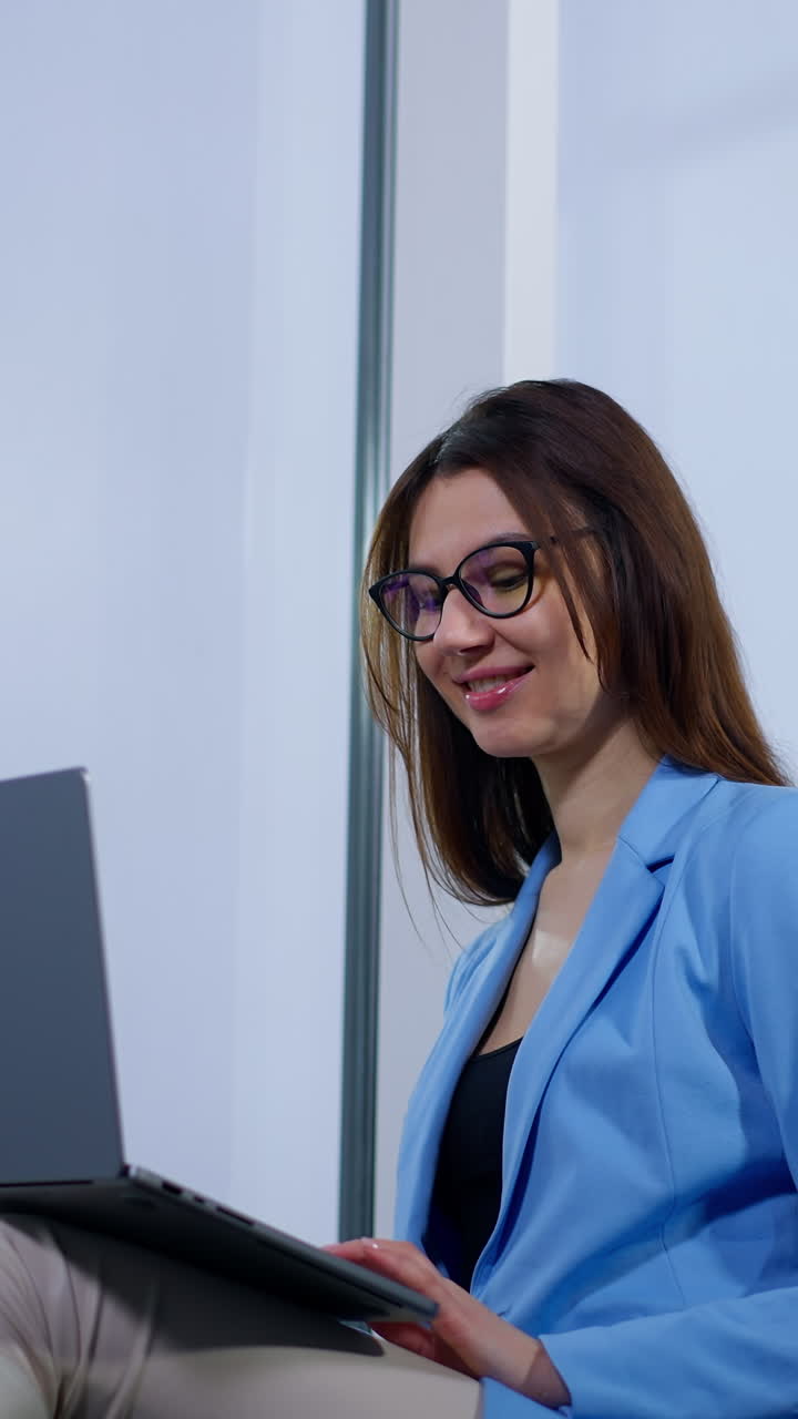 Lady in glasses is busy with her work at laptop. Woman using gadget keeping it on her laps. Low angle view. Vertical video