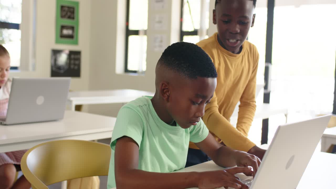 African American youth males typing on silver laptop in class as standing peer approaching, guiding
