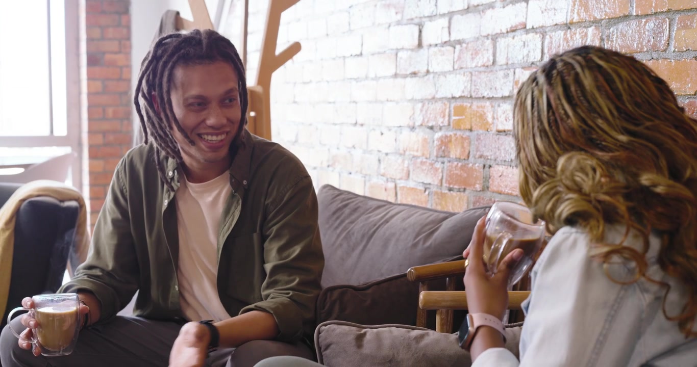 African friends sitting sofa in loft, man starting chat, gesturing while woman sipping cups