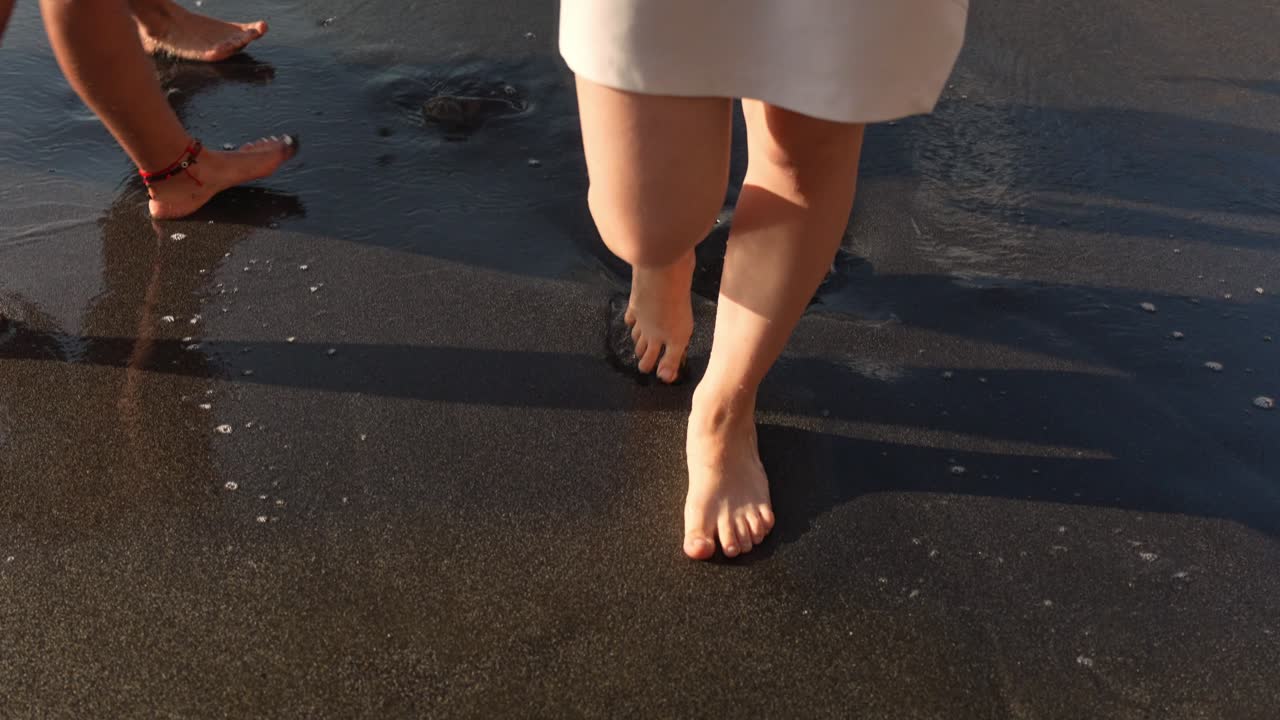 pies femeninos caminan por la playa de arena negra, envueltos por pequeñas olas blancas